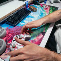 Overhead view of a gamer's hands on a controller at a desk with a mechanical keyboard, headphones, and a Gamer Grip for Gaming tube and box