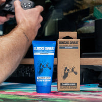 Blue Gamer Grip for Gaming tube and kraft box on a desk mat, with a gamer holding a controller in the background