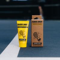 Yellow Gamer Grip for Racquet & Paddle tube and kraft box standing on the line of a blue tennis and pickleball court